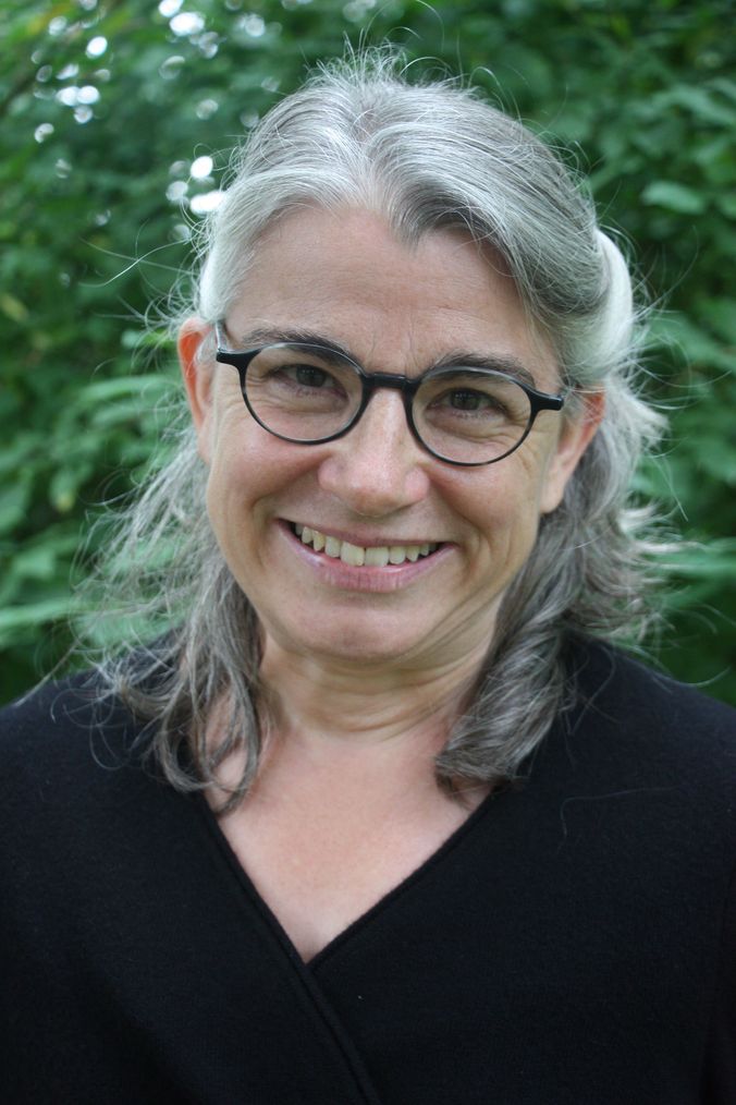 Lächelnde Person mit grauen Haaren und Brille vor grünem Hintergrund.