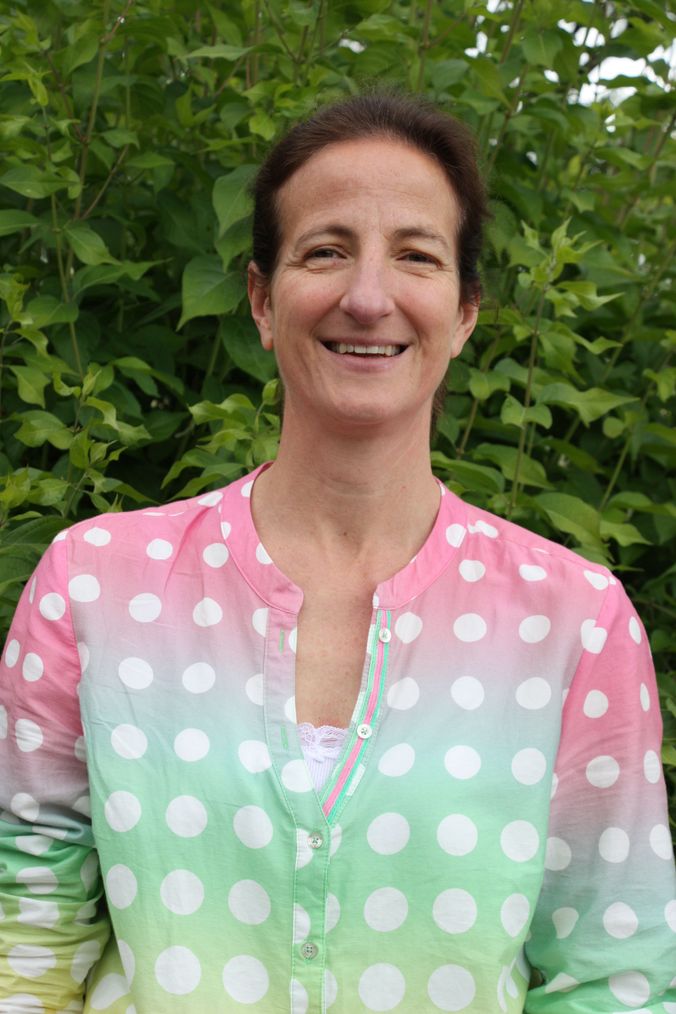 Lächelnde Frau in pink-grünem Polka-Dot-Shirt vor grünem Blattwerk.