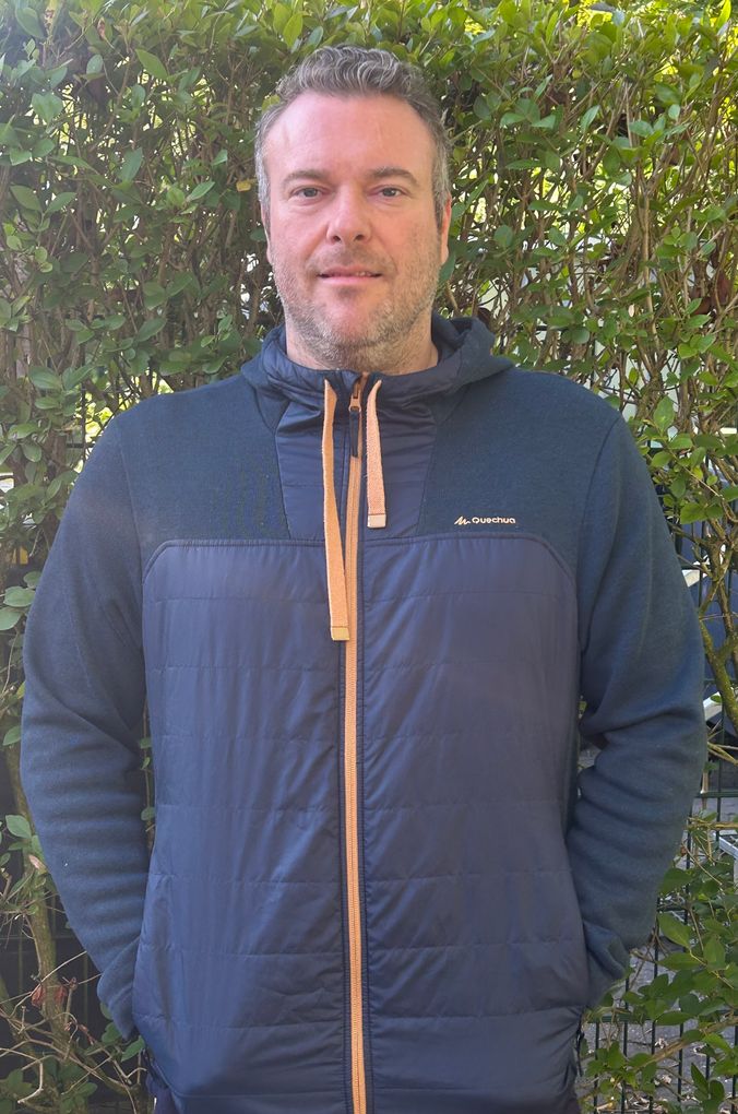 Mann trägt dunkelblauen Softshell-Jackett mit Zip-Front vor grünem Buschwerk.