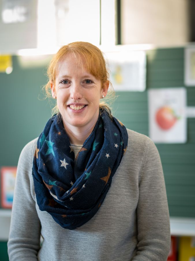 Lächelnde Person mit grauer Jacke und bunter Sternen-Schal vor Klassenzimmer.