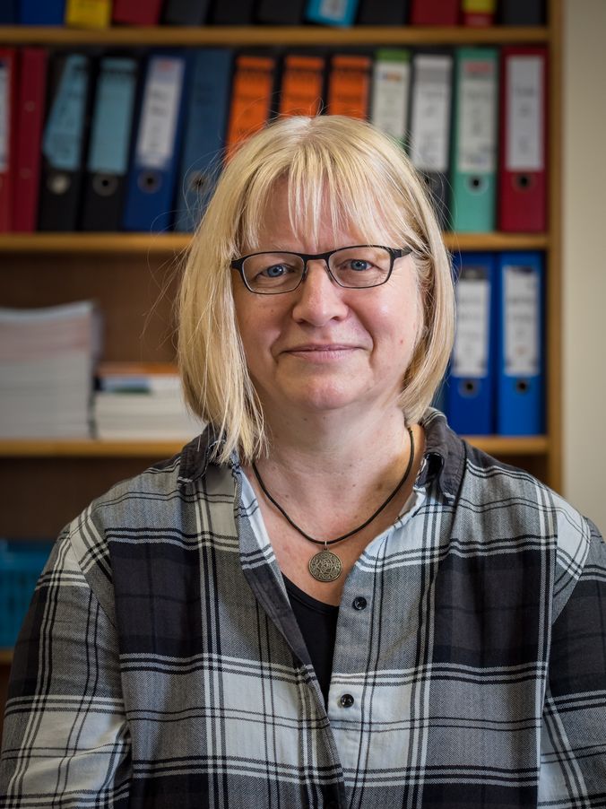Frau mit Brille und Plaid-Shirt vor Regal mit Ordnern lächelnd.