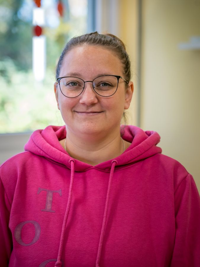 Lächelnde Person mit Brille in pinkem Hoodie vor hellem Hintergrund.