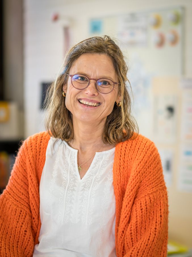 Lächelnde Frau mit Brille in orangefarbenem Pullover vor buntem Hintergrund.