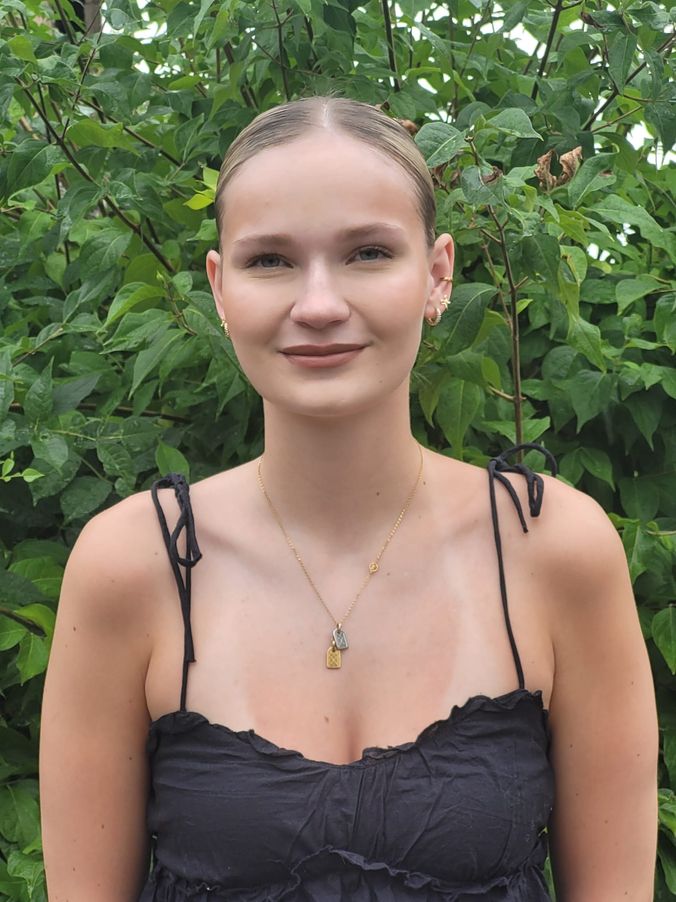 Frau lächelt vor grünem Blattwerk in schwarzem Kleid mit dünnen Trägern und goldener Kette.