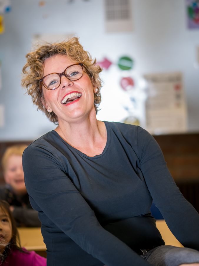 Glückliche Frau mit Brille lacht in einem bunten Klassenzimmer.
