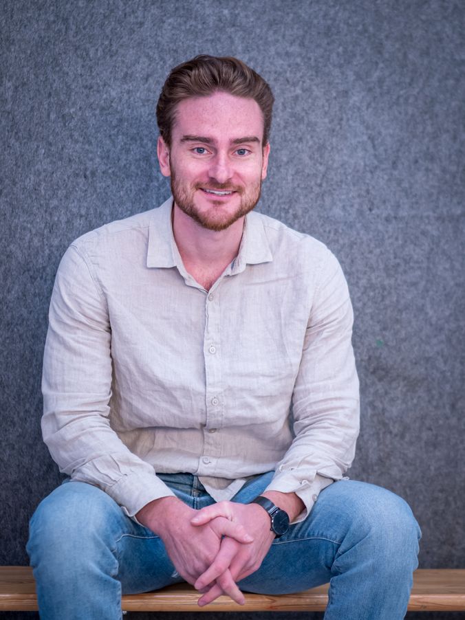 Lächelnder Mann in hellem Hemd und Jeans sitzt entspannt auf Stuhl vor grauer Wand.