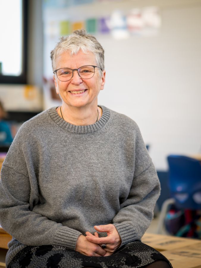 Lächelnde Person mit Brille trägt grauen Pullover und sitzt in einem Klassenzimmer.
