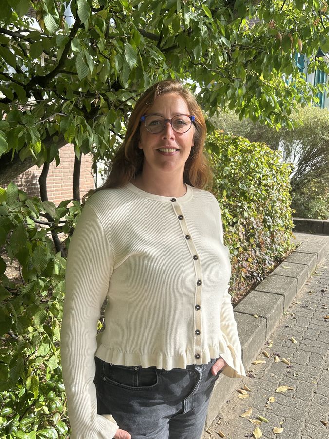 Frau mit roten Haaren lächelt im Freien in cremefarbenem Pullover und Jeans.
