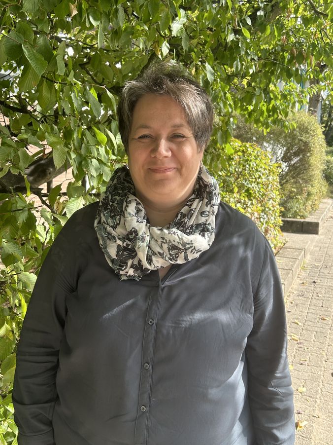 Frau mit Schal lächelt vor grünem Blattwerk im Freien.