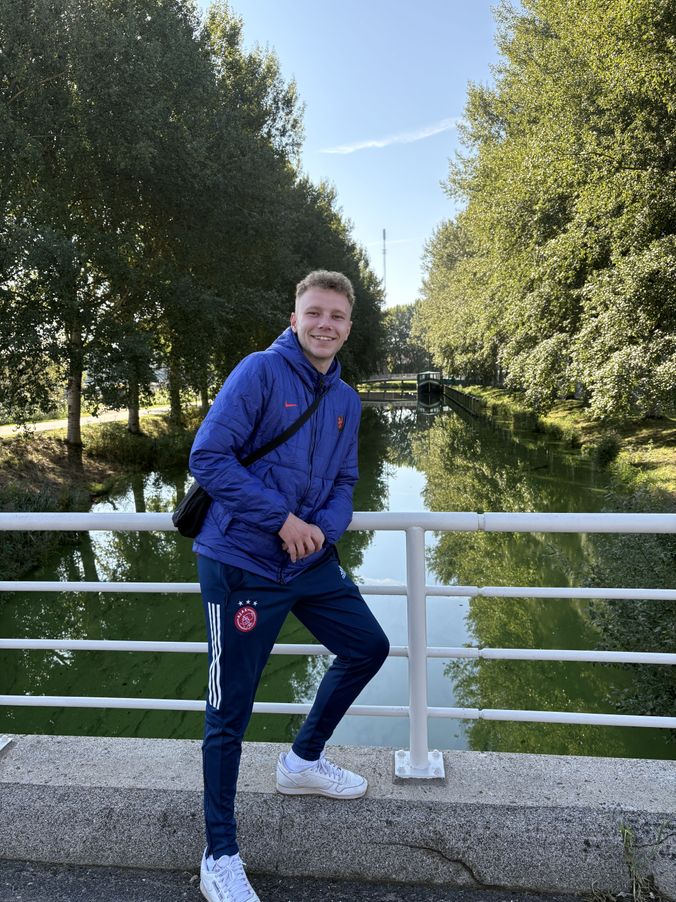 Junger Mann in blauem Adidas-Jackenzug lässig an Geländer mit Wasserlauf im Hintergrund.
