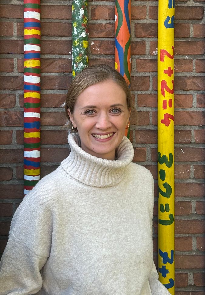 Lächelnde Frau vor bunt bemalten Pfosten und Backsteinmauer, cremefarbenes Turtleneck-Shirt.